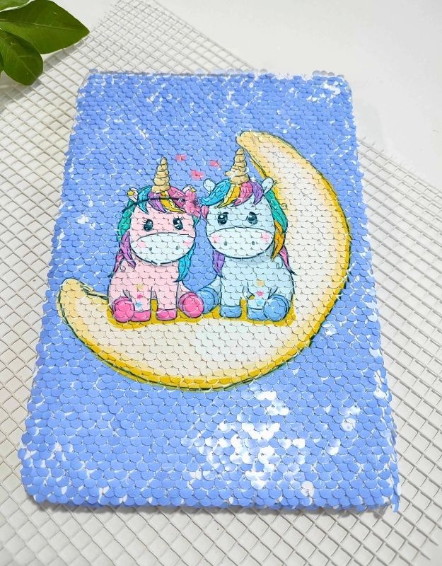 Sequined notebook with cartoon unicorns on a moon, on a textured surface.